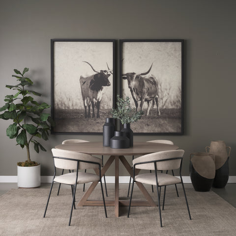 Dining room with a round wooden table and chairs, framed black and white pictures of cows on the wall, and a plant.